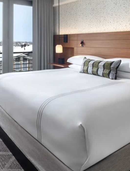 Modern hotel room with a large neatly made bed, a desk and chair, and glass doors leading to a balcony with a snowy view of wooden buildings outside.