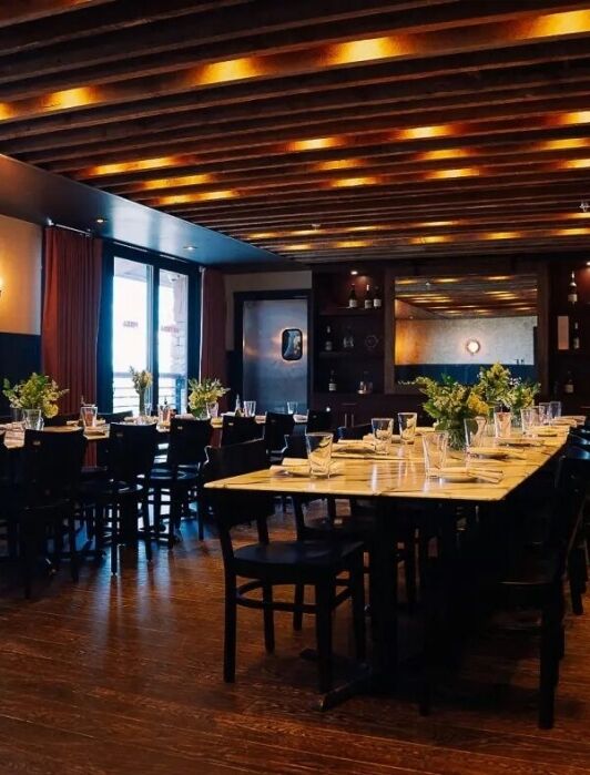 A modern dining room with two long tables set for a meal, surrounded by chairs, wooden floors, ceiling beams, and warm lighting.