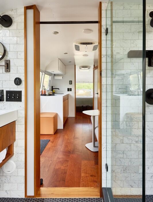 Modern bathroom with white tile walls, wooden vanity, and glass shower in an Airstream trailer, looking into the kitchen and dining area with wood floors and natural light.