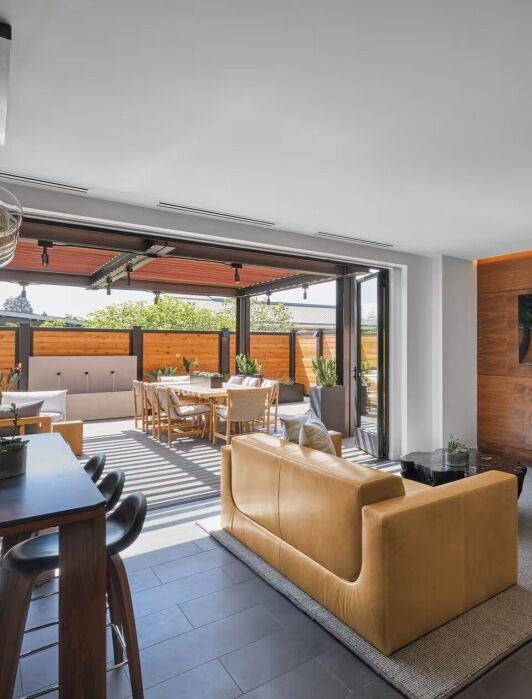 Modern open-concept living space with a tan sofa, wall-mounted TV, dining area, and outdoor patio visible through large sliding glass doors.