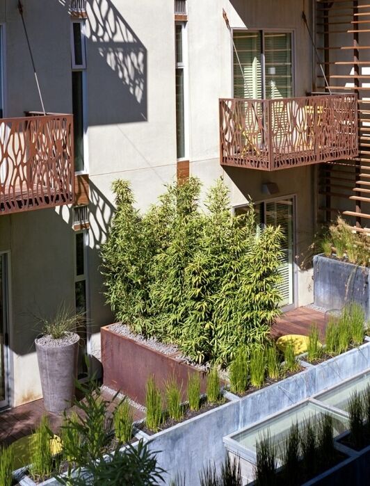 Modern apartment building with decorative metal balconies, large windows with blinds, potted plants, and lush greenery on the terrace.