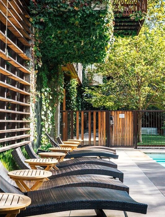 Wicker lounge chairs and small tables line the edge of an outdoor pool, surrounded by greenery and wooden fencing on a sunny day.