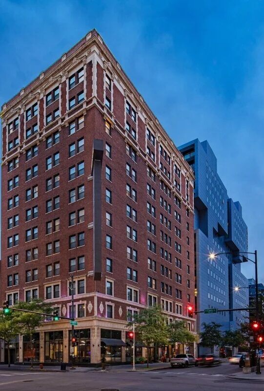 A multi-story red brick building with white trim stands on a city street corner at dusk, with modern high-rises and streetlights visible in the background.