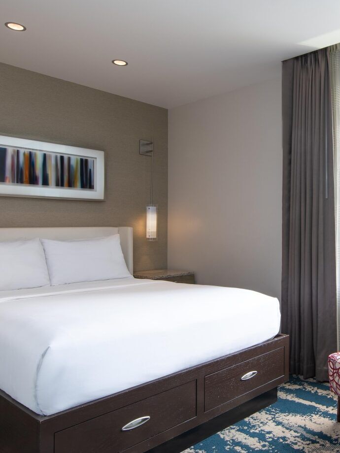 Hotel room with a neatly made bed, side tables, a patterned chair, a desk with a lamp, and a window showing city buildings in daylight.