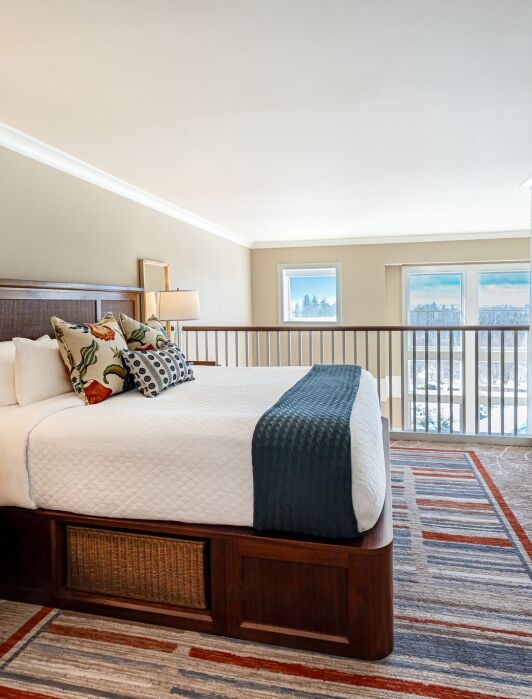 A hotel room with a neatly made bed, bedside tables with lamps, a wall-mounted TV, large windows, and a balcony overlooking a scenic view.