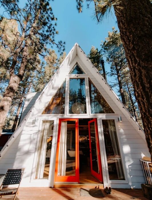 A white A-frame cabin with large windows and open red double doors, surrounded by tall pine trees under a clear blue sky.