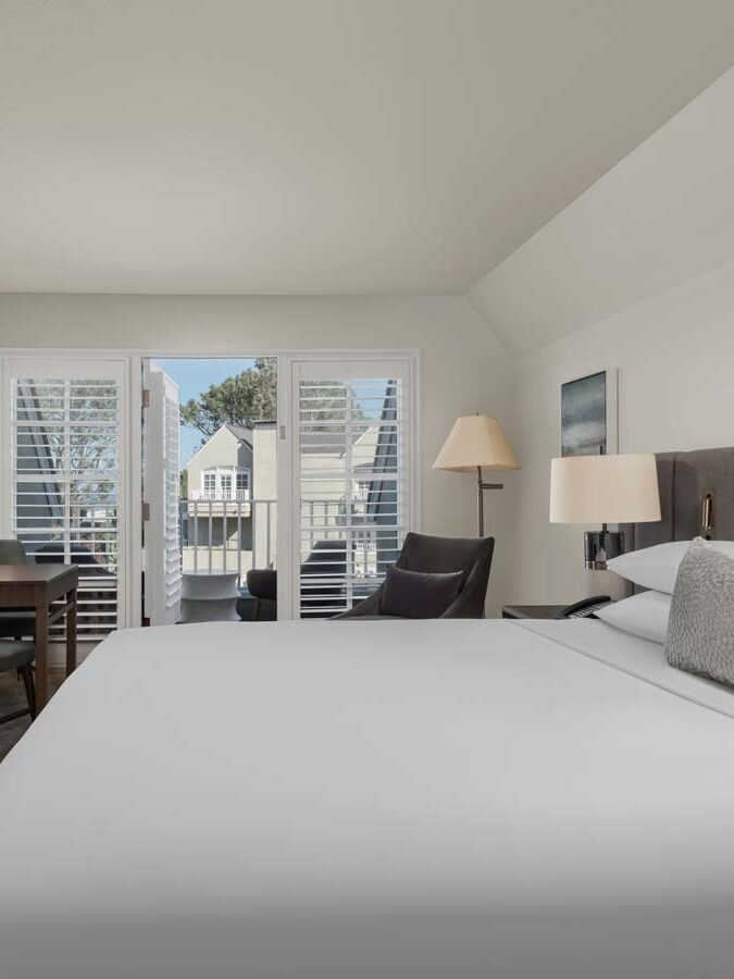 A hotel room with a large bed, fireplace, TV, desk, armchair, and balcony doors overlooking houses and trees.