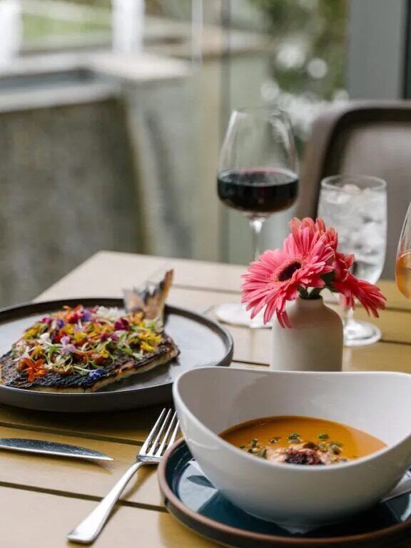 A table set with a bowl of soup, a plate of fish topped with herbs, a glass of red wine, a glass of white wine, water with ice, and a pink flower in a vase.