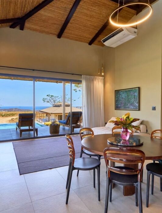 Modern living and dining area with round table, chairs, sofa, and large windows opening to a patio with pool and ocean view; bedroom visible through open doorway.