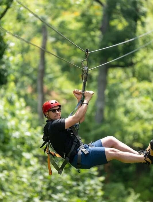 A person wearing a red helmet, sunglasses, and harness rides a zipline through a green forest on a sunny day.