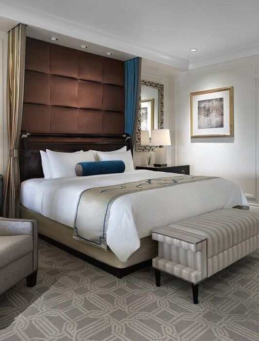 Modern hotel room with a large bed, upholstered headboard, armchair, striped bench, two lamps, and neutral-toned decor.