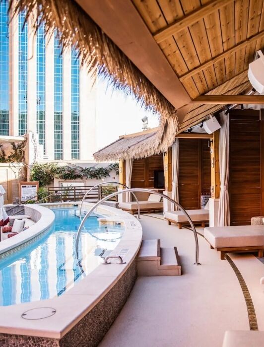 Poolside cabanas with wooden walls and lounge seating are shown next to a curved outdoor swimming pool surrounded by modern buildings.