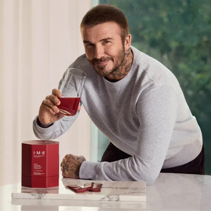 A man with tattoos in a grey sweatshirt holds a glass of red beverage, leaning on a counter next to a red box labeled "IM-8" and red packets.