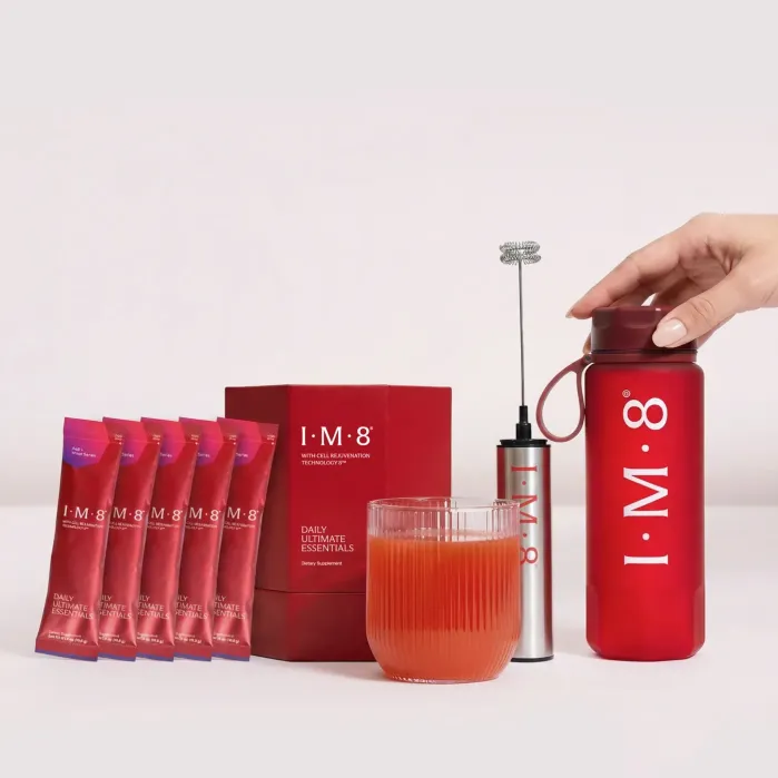 A hand holds a red shaker bottle labeled "IM·8" next to supplement packets, a box, a glass of red drink, and a small handheld frother, all on a white background.