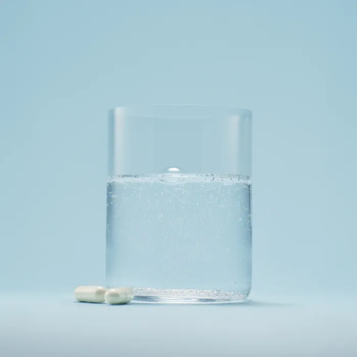 A clear glass of sparkling water with two white capsules placed beside it on a light blue background.