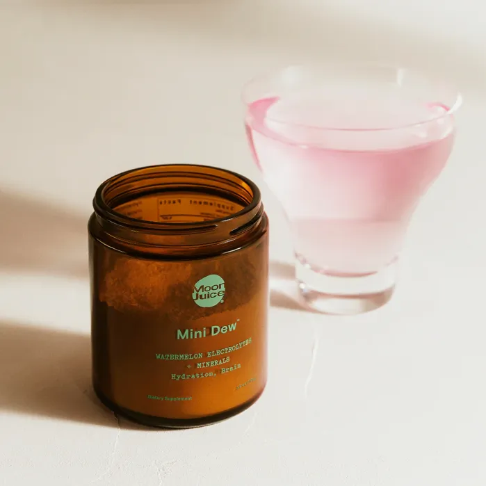 A brown jar labeled “Mini Dew” by Moon Juice sits next to a glass of pink liquid on a light-colored surface.
