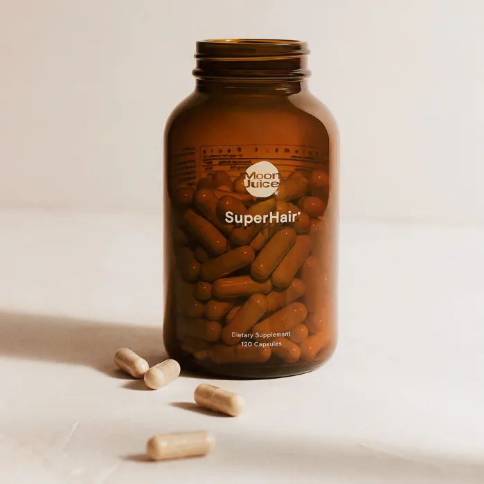 A brown bottle labeled "SuperHair" by Moon Juice, containing capsules, with four capsules lying on a light surface in front of the bottle.