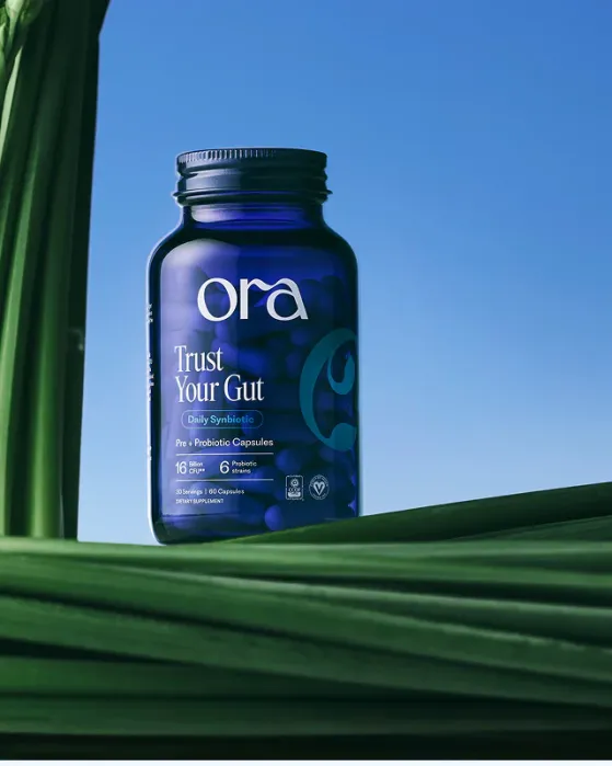 A blue bottle of Ora "Trust Your Gut" pre and probiotic capsules sits on green palm leaves against a clear blue sky.