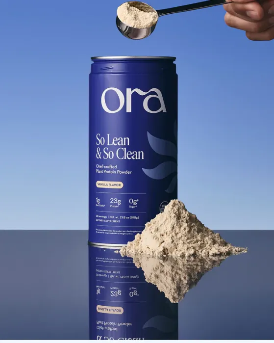 A hand holds a scoop of vanilla plant protein powder above an Ora So Lean & So Clean container, with a pile of powder and its reflection on the surface below.