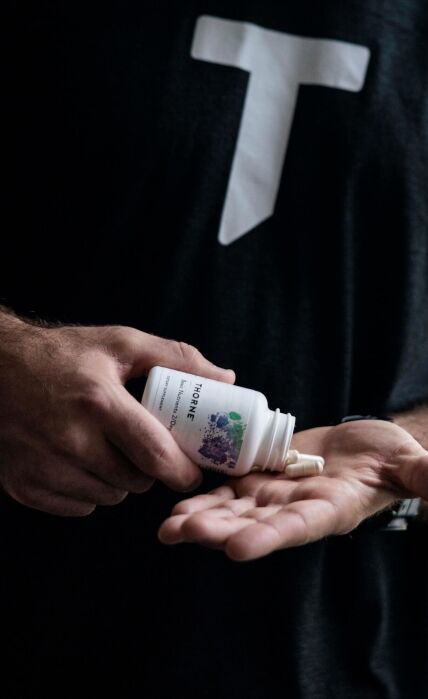 A person pours capsules from a white supplement bottle into their hand, wearing a black shirt with a large white "T" printed on it.