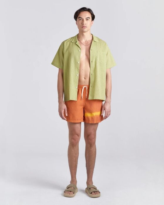 A man stands facing forward wearing a light green short-sleeve shirt, orange tie-dye swim shorts, and beige sandals against a plain light background.