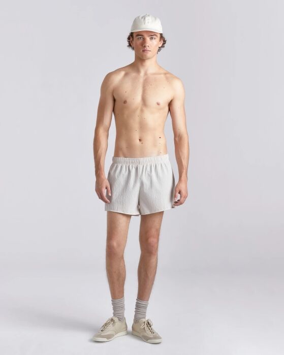 A young man stands facing forward, wearing a white cap, white shorts, gray socks, and beige sneakers against a plain light background.