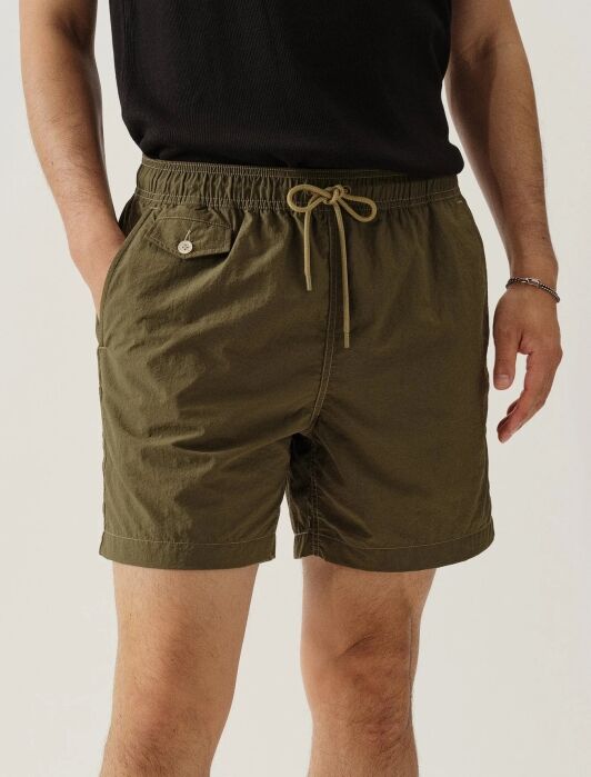 A person wearing olive green drawstring shorts with a side pocket and a black t-shirt, standing with hands in pockets.