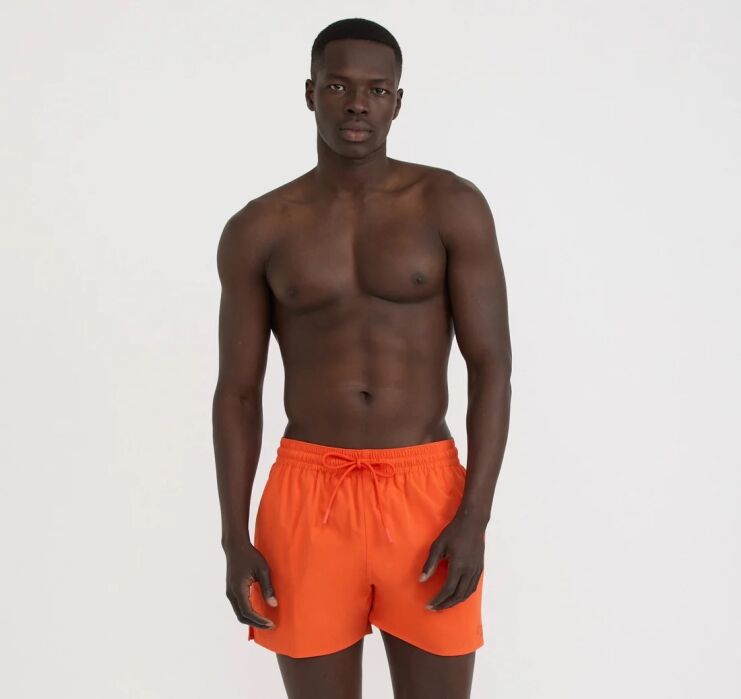 A man stands against a plain white background wearing bright orange swim trunks; he is shirtless and facing forward.