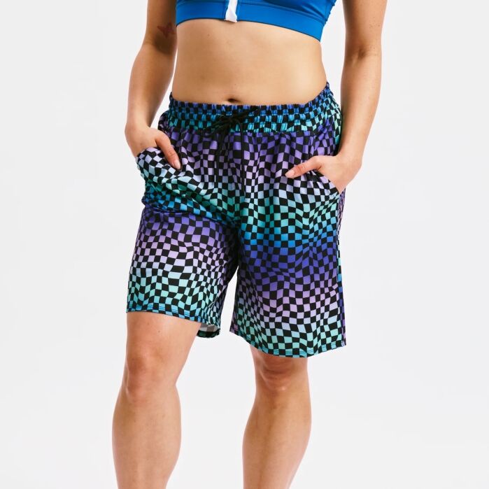 Person wearing a blue sports bra and checkered shorts with a black, teal, and purple geometric pattern, standing with hands in pockets against a plain white background.