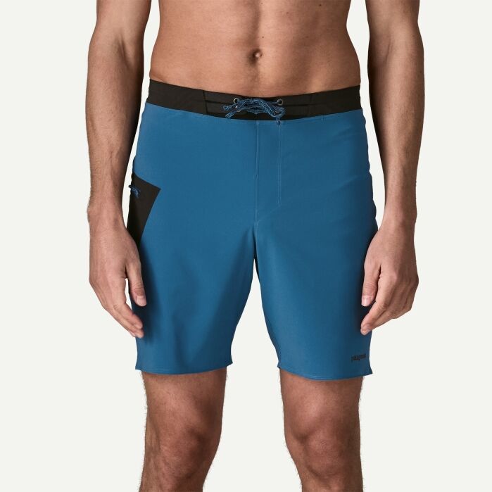 A man wearing blue and black board shorts stands facing forward; his torso and upper legs are visible, with arms relaxed at his sides.