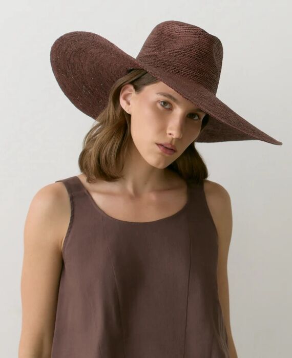 Woman wearing a wide-brimmed brown hat and a sleeveless brown top, standing against a plain light background, looking directly at the camera.