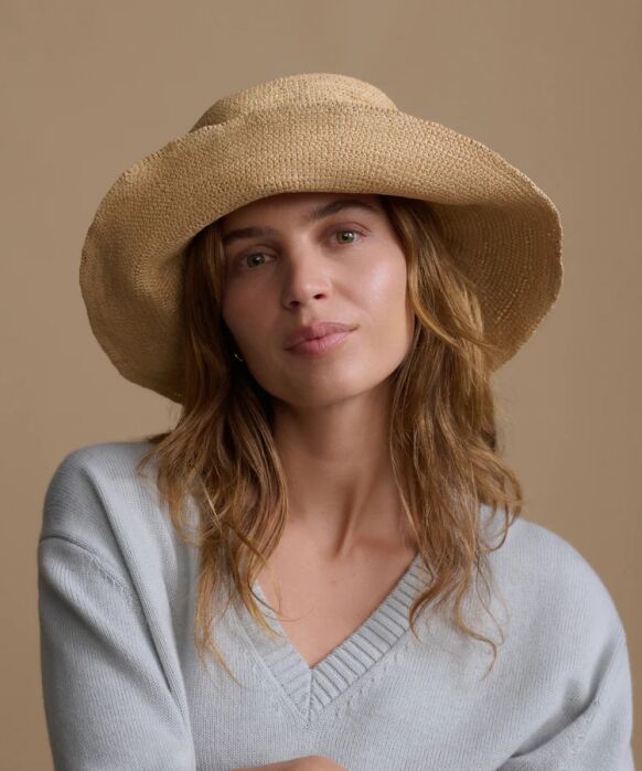A woman with wavy light brown hair wears a wide-brimmed straw hat and a light gray v-neck sweater, posing against a plain beige background.