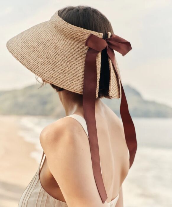A woman in a sleeveless dress stands on a beach, facing away. She wears a wide-brimmed straw visor hat with a large brown ribbon tied at the back.