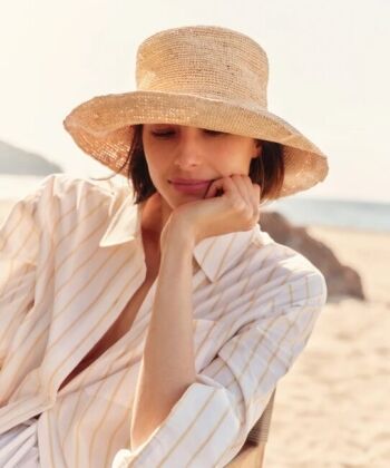 Woman sitting on a sandy beach wearing a wide-brimmed straw hat and a light-colored striped shirt, resting her chin on her hand.