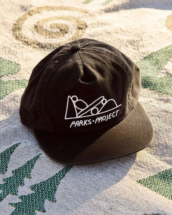 A black baseball cap with a white “PARKS PROJECT” logo sits on a patterned fabric surface in sunlight.