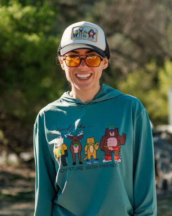 Woman wearing sunglasses, a hat, and a blue hoodie with cartoon animals and the words “Adventure With Friends,” smiling outdoors.
