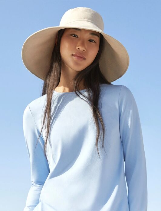 Woman wearing a wide-brimmed beige hat and light blue long-sleeve shirt stands outdoors against a clear blue sky.