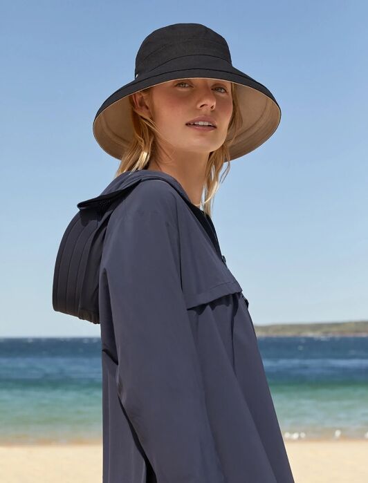 Woman wearing a wide-brimmed black hat and navy jacket stands on a sandy beach with blue water and a clear sky in the background.