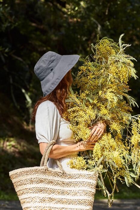 Person wearing a gray hat and light dress holds a large bouquet of yellow flowers and carries a woven basket outdoors.