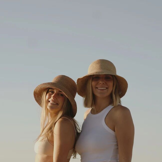 Two women wearing sun hats and light tops stand outdoors, smiling at the camera against a clear sky.