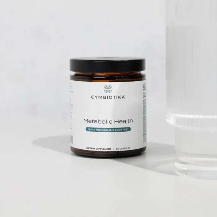 A jar of Cymbiotika Metabolic Health dietary supplement capsules is placed next to a clear glass of water on a light surface.