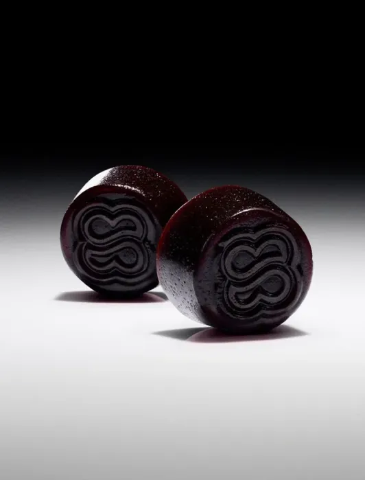 Two round, dark-colored candies with a shiny surface and embossed swirl patterns are positioned on a white surface against a dark gradient background.