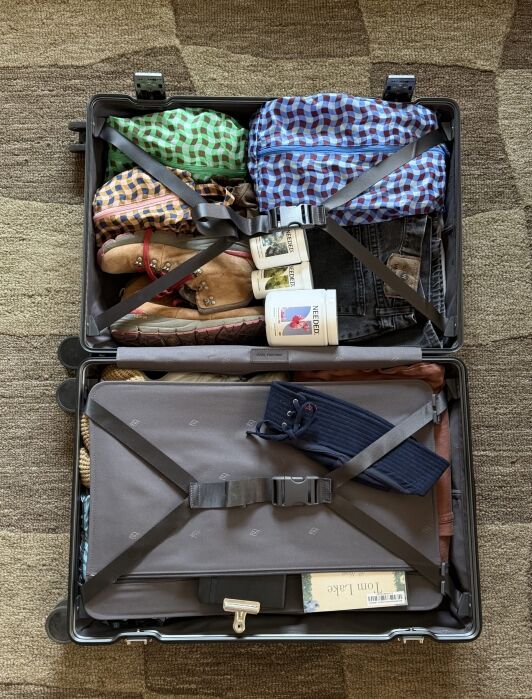 Open suitcase neatly packed with folded clothes, shoes, toiletries, a book, socks, and a laptop, all secured with suitcase straps on a patterned carpet.