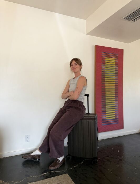 Person in a sleeveless top and loose pants sits on a suitcase in a room with a colorful rectangular wall art and dark floor.