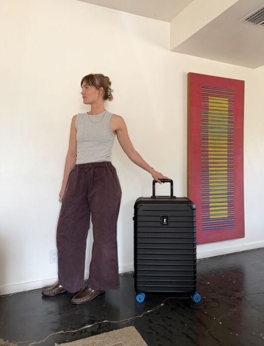 A person in a gray tank top and loose brown pants stands indoors holding a black rolling suitcase next to a wall with colorful abstract artwork.