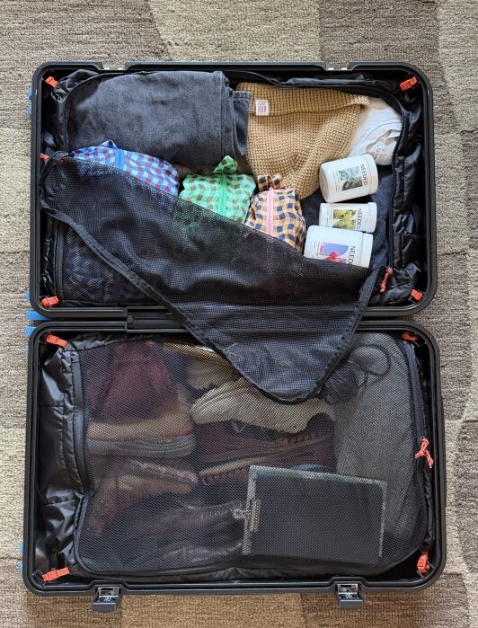 Open suitcase containing folded clothes, several bottles, and a tablet, organized under mesh compartments on a patterned carpet.