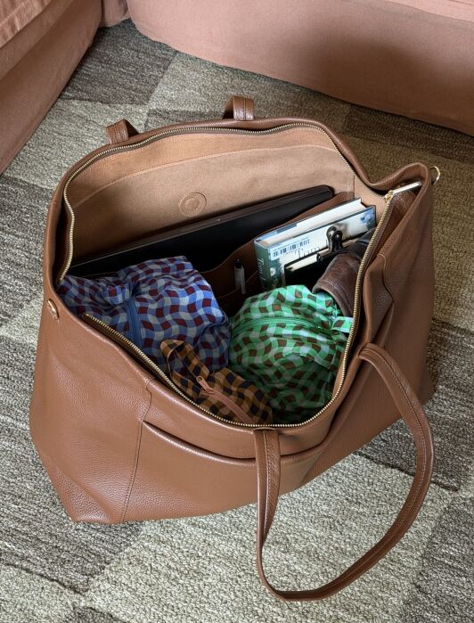 Open brown leather tote bag on a carpet, containing colorful folded shirts, a laptop, books, and a wallet.