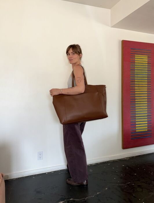 A woman stands indoors holding a large brown leather tote bag, wearing a gray tank top and dark pants, with a colorful artwork on the wall behind her.