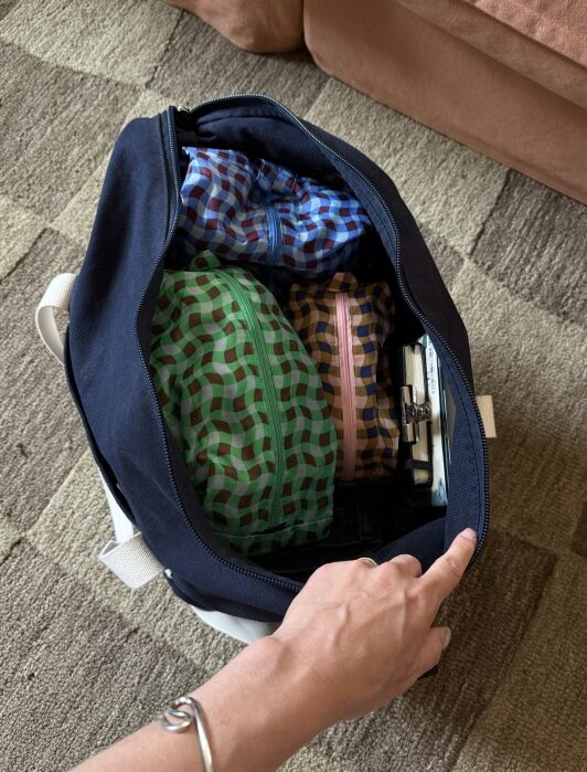 A hand holds open a navy bag containing three patterned pouches and a phone placed side by side on a patterned carpet.