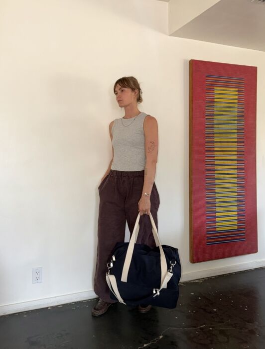 A woman in a gray tank top and loose brown pants stands indoors holding a large navy bag, with a red and yellow striped artwork on the wall behind her.